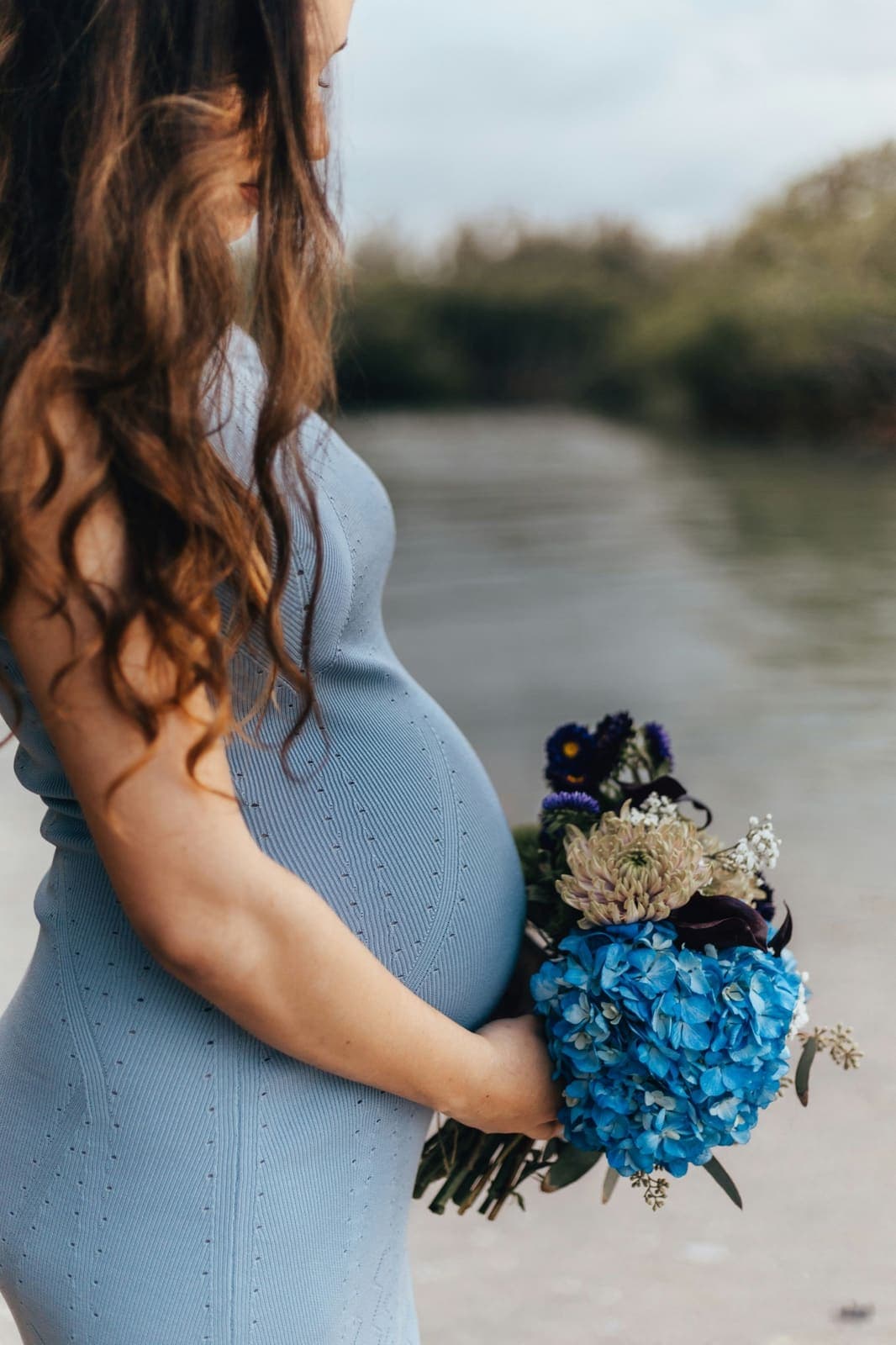 Schwangere in blauem Kleid hält einen Blumenstrauß am Wasser